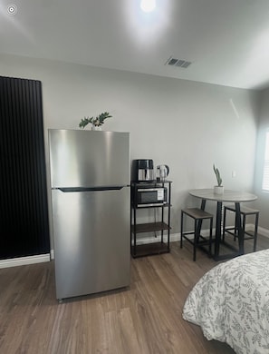 A compact hotel room with a stainless steel refrigerator, a coffee maker, a small table with two stools, a potted plant, and a bed with a patterned bedspread. - Serene Brand New studio with AC in charming La Palma  (La Palma)