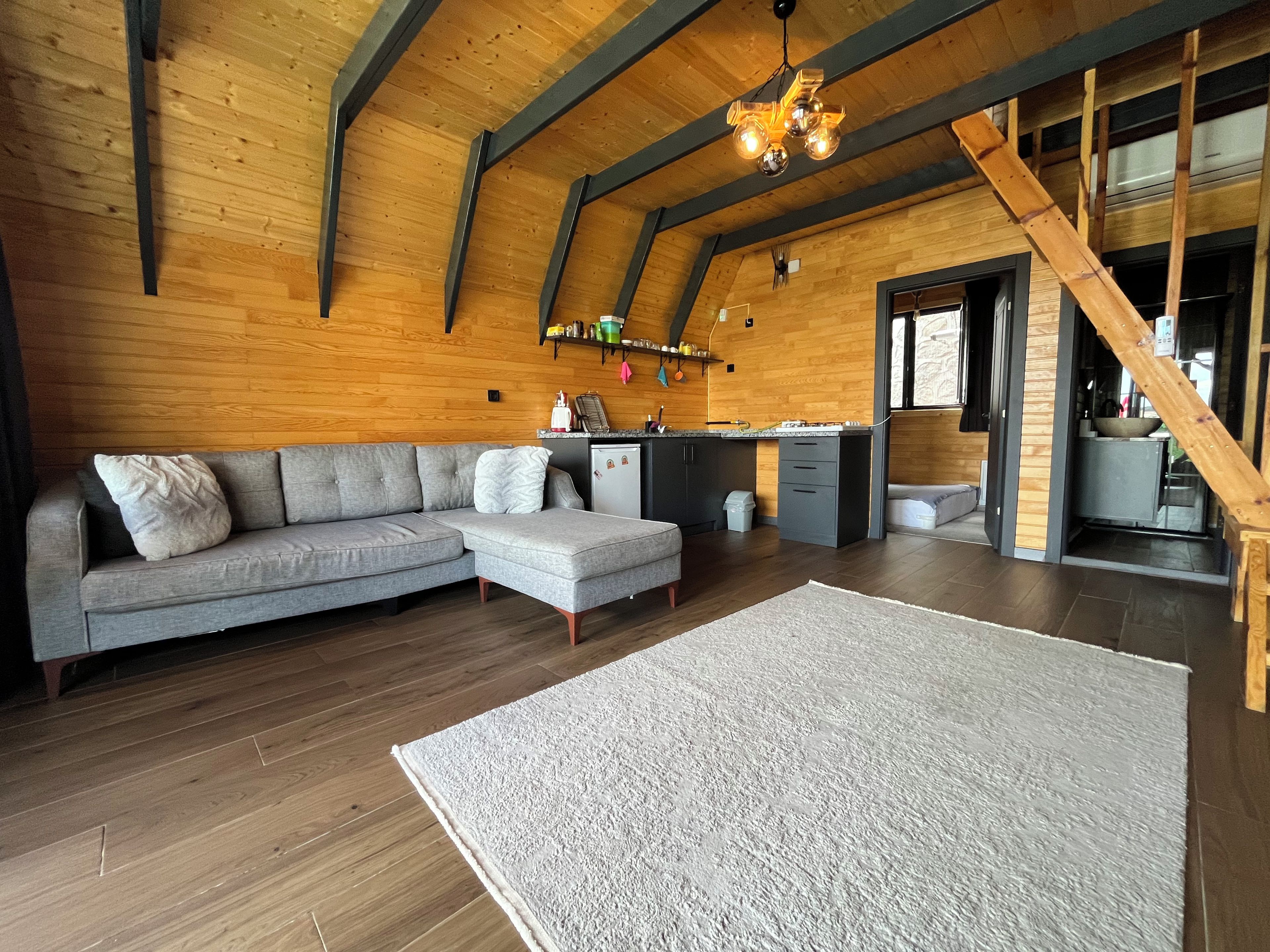 Ein modernes Interieur mit einem grauen Ecksofa, einem weißen Teppich, Holzböden, einer Küchenzeile und einer Treppe.