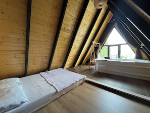 Una habitación con techo inclinado, paredes de madera, una cama con colchón y almohada blancos, y una ventana que ofrece vistas del exterior.