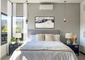 Ein modernes Schlafzimmer mit einem großen Bett, zwei Nachttischen mit Lampen, einem Fenster mit Vorhängen und einem Bild an der Wand.