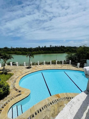 A swimming pool with a curved edge, surrounded by a white fence, and a view of a natural landscape in the background. - Mango Villa (Mtwapa)