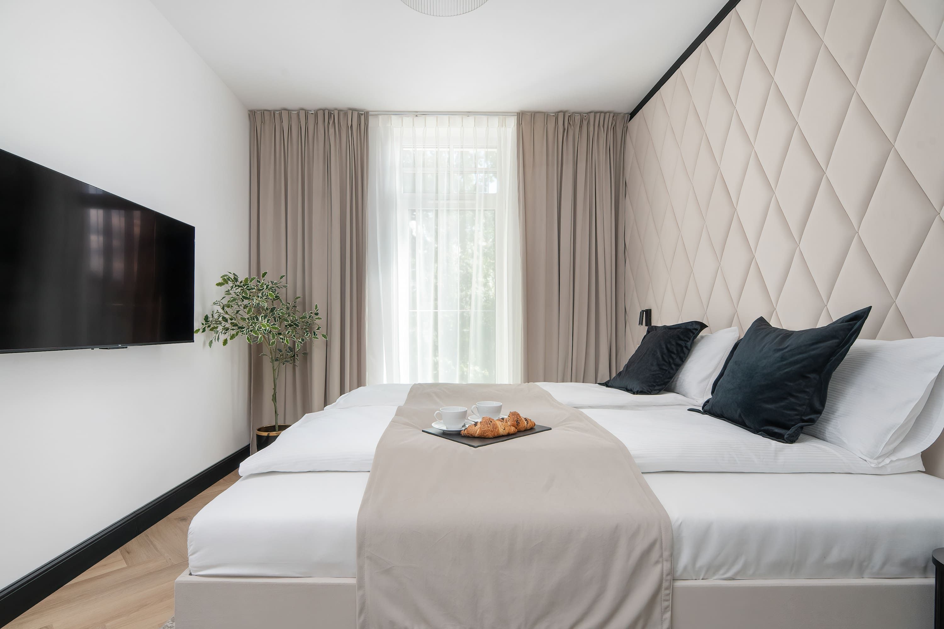 A hotel room with a bed, a TV mounted on the wall, a potted plant, and a window with curtains.