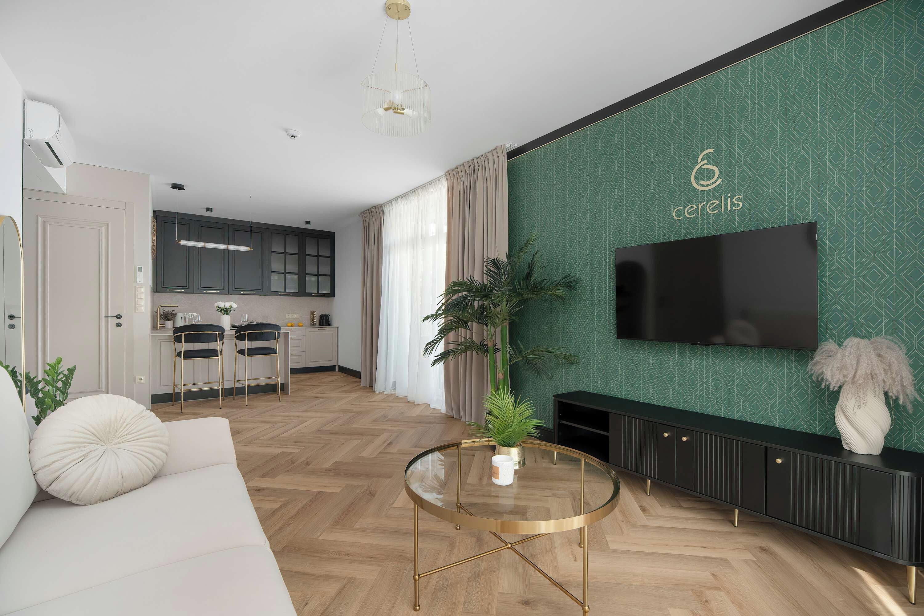 Ein modernes Wohnzimmer mit einer Couch, einem Glastisch, einem an einer grün gemusterten Wand montierten Fernseher und einer Küche im Hintergrund.