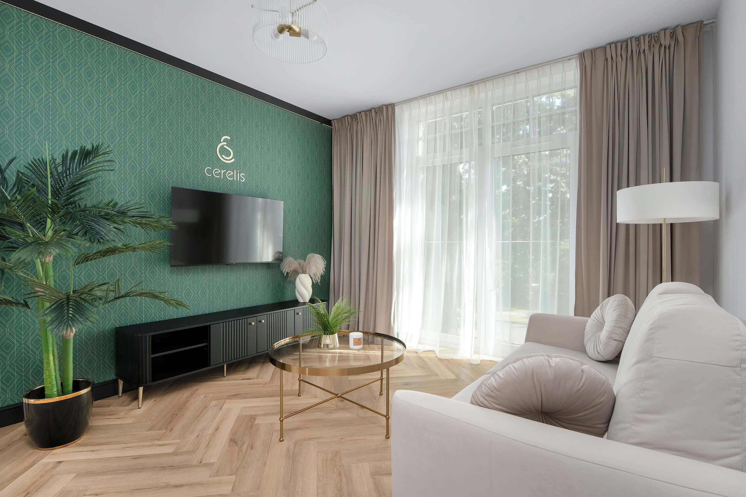 A modern living room with a green patterned wall, a flat-screen TV, a sofa, a coffee table, and a large window with curtains.