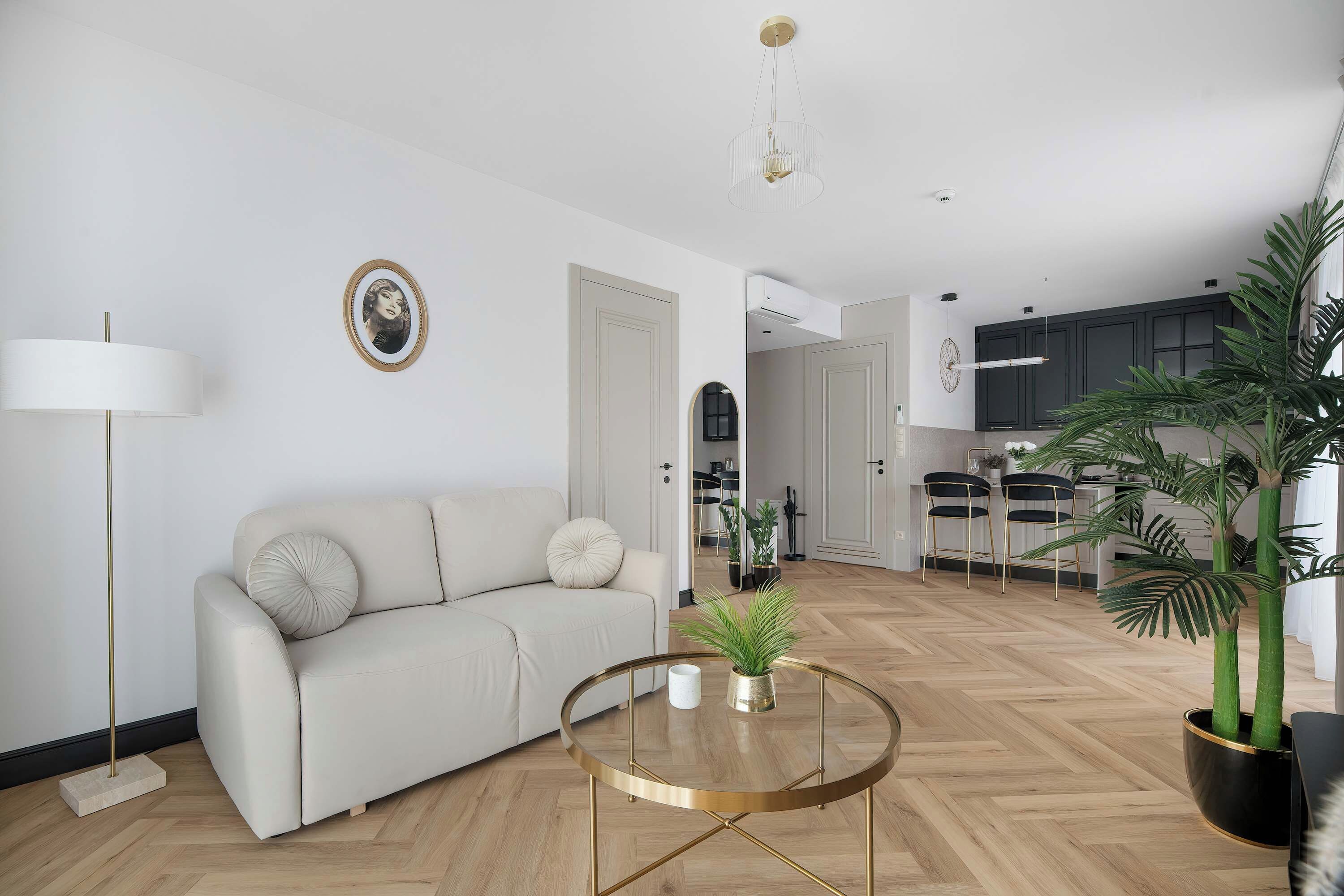 A modern living room with a sofa, a round coffee table, and a floor lamp. There is a kitchen in the background with dark cabinets and a hanging light fixture.