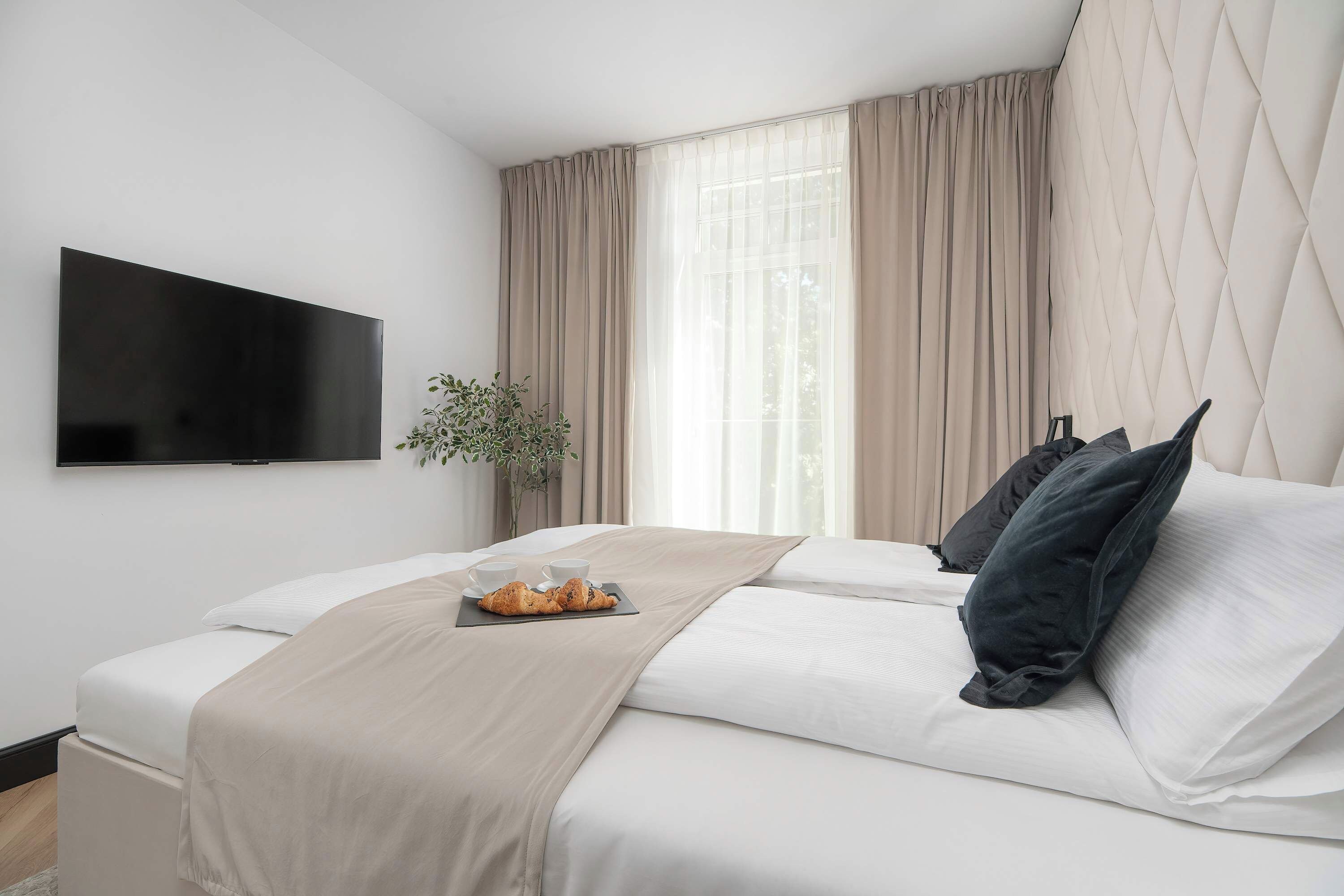 Ein modernes Hotelzimmer mit einem flachbildfernseher an der Wand, einem Bett mit weißen Leinen, einer beige farbenen Tagesdecke und einem Tablett mit Croissants.