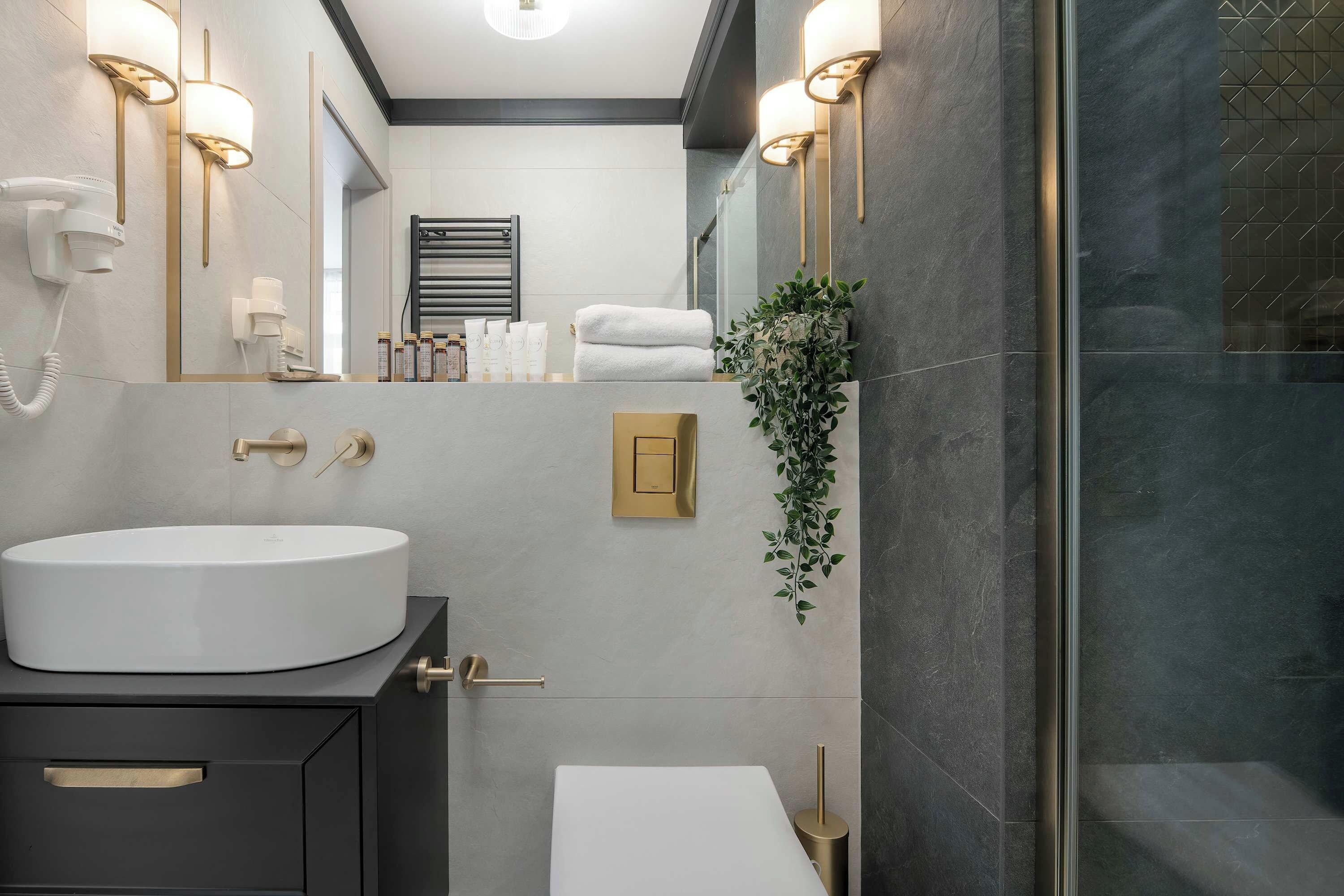 Ein modernes Badezimmer mit einem großen weißen Waschbecken, goldenen Armaturen, einem Handtuchhalter mit Handtüchern und einer Topfpflanze.