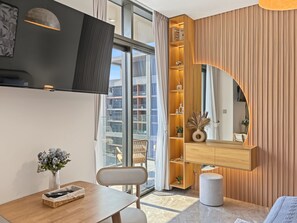 A modern interior with a dining area, a built-in wooden shelf, and a large window with a view of buildings. - Peninsula Two - 3410 Studio (Dubai)