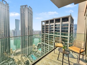 A balcony with a glass railing, two wicker chairs, a small table, and a view of a cityscape with tall buildings and a body of water. - Peninsula Two - 3410 Studio (Dubai)