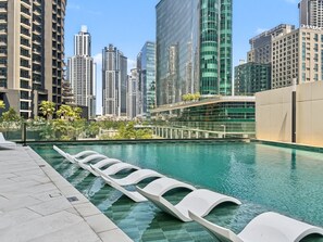 A modern swimming pool with lounge chairs, surrounded by high-rise buildings and a glass structure. - Peninsula Two - 3410 Studio (Dubai)