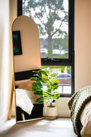 A room with a bed, a potted plant, and a mirror reflecting a window view. - Seednest Modern Apt by Mall, BBQ & Parking (Doncaster East)