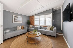 Ein Wohnzimmer mit grauen Sofas, einem Holztisch, einem großen Fenster und einer Ziegelwand.