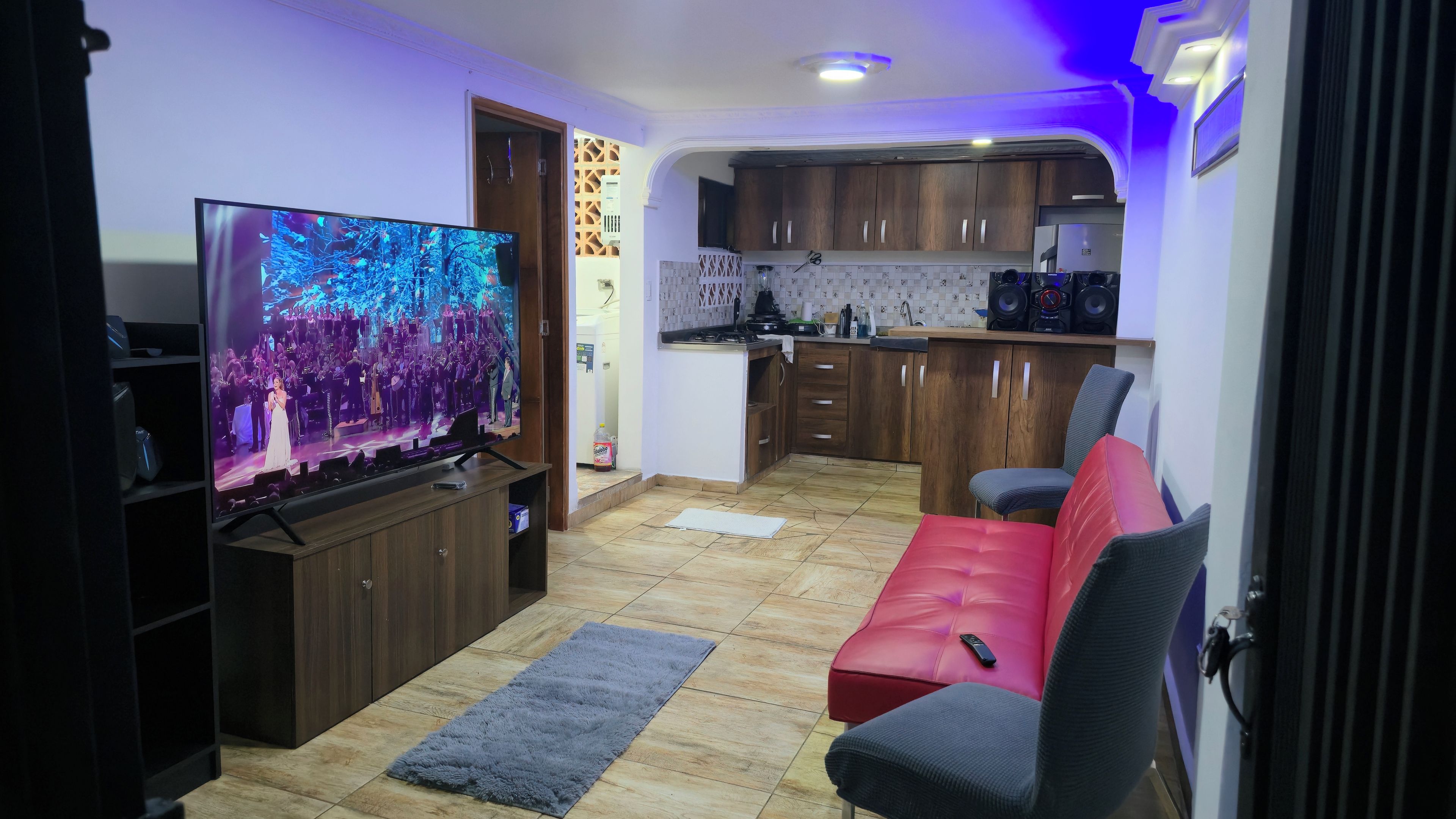 A living room with a flat-screen TV, a red sofa, a kitchen area with wooden cabinets, and a tiled floor.