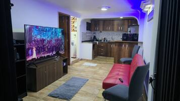 A living room with a flat-screen TV, a red sofa, a kitchen area with wooden cabinets, and a tiled floor.
