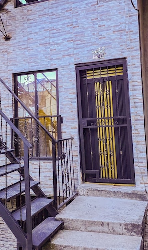 Un edificio con fachada de ladrillo, una puerta negra con franjas verticales amarillas y una escalera que lleva a un nivel superior.