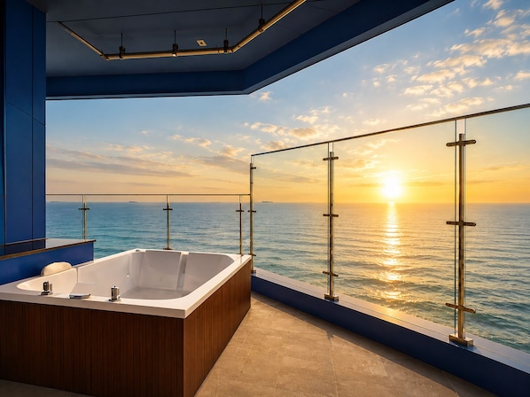 Un baño moderno con una bañera grande, un tocador de madera y un balcón acristalado con vistas al mar al atardecer.
