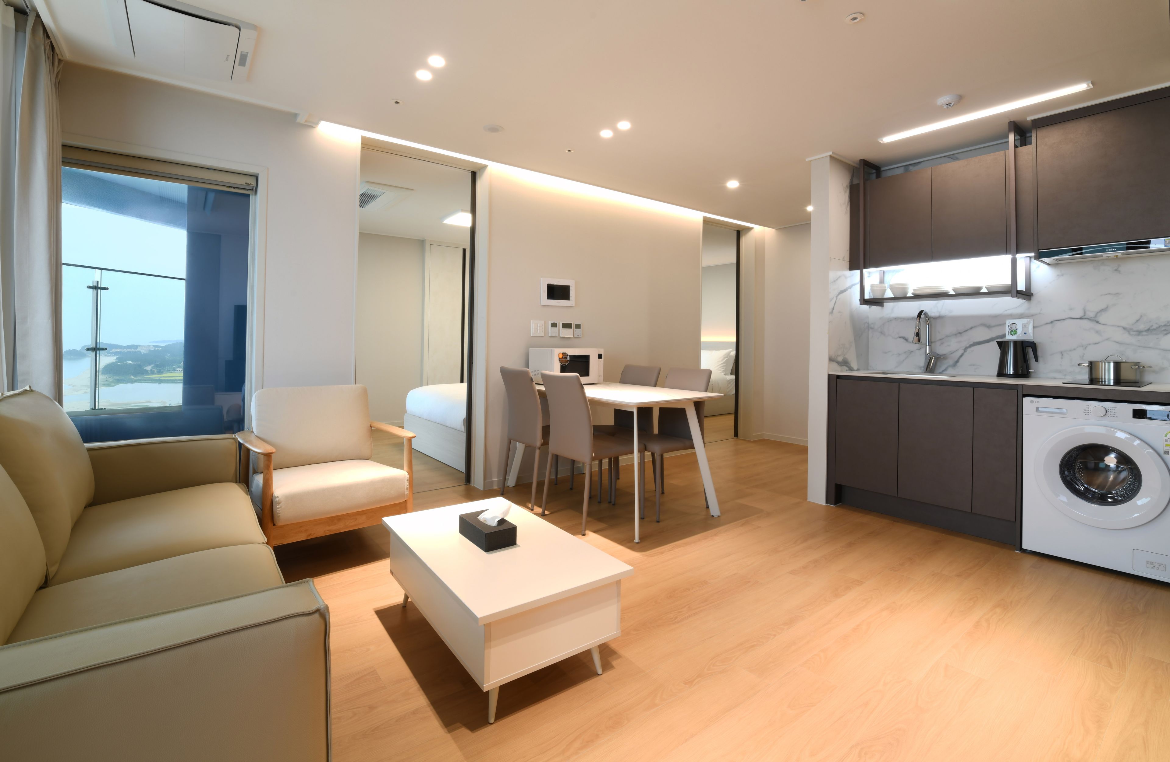Una habitación de hotel moderna con un sofá, una mesa de comedor con sillas, una zona de cocina con lavadora y un amplio ventanal con vistas.