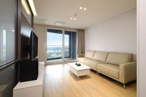 A modern living room with a large flat-screen TV, a beige sofa, a white coffee table, and a large sliding glass door leading to a balcony with a city view.