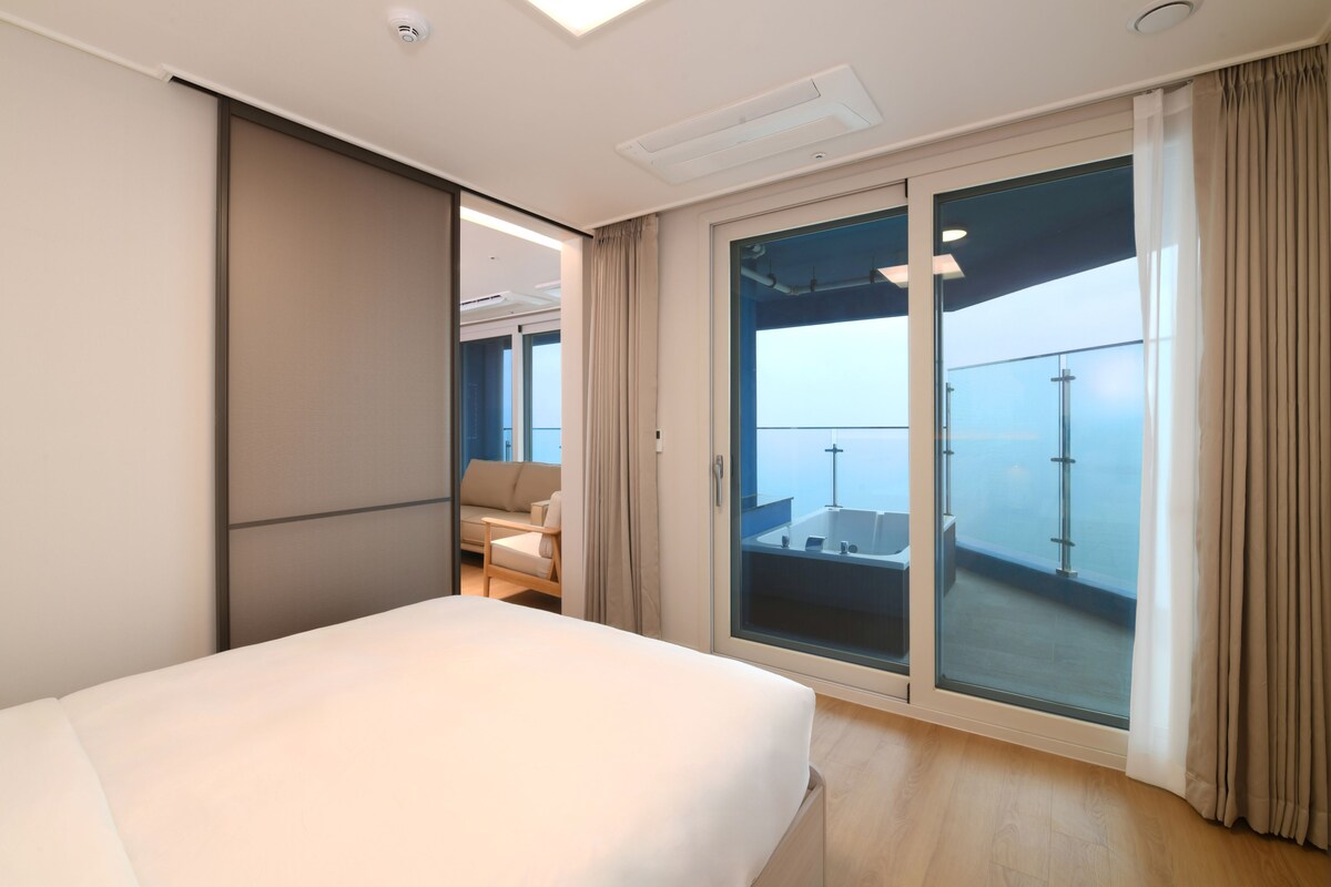 Een moderne hotelkamer met een groot bed, een zithoek met een bank en fauteuils, en een glazen schuifdeur die leidt naar een balkon met uitzicht.