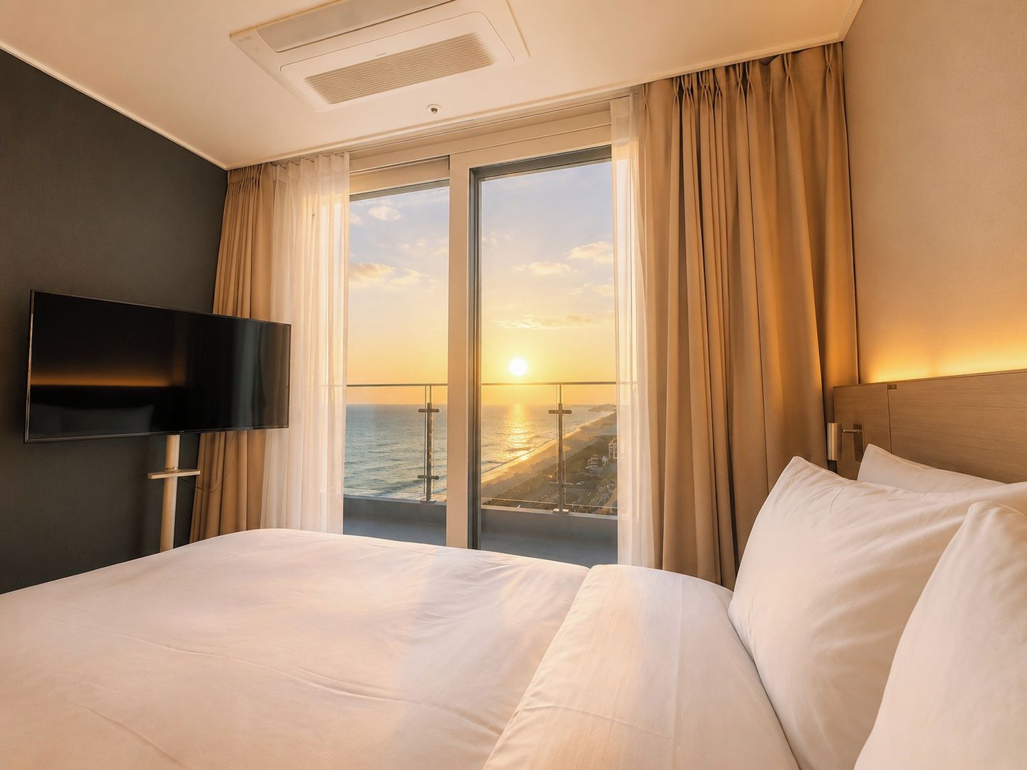 A hotel room with a bed, a television, a large window with curtains, and a view of the ocean.