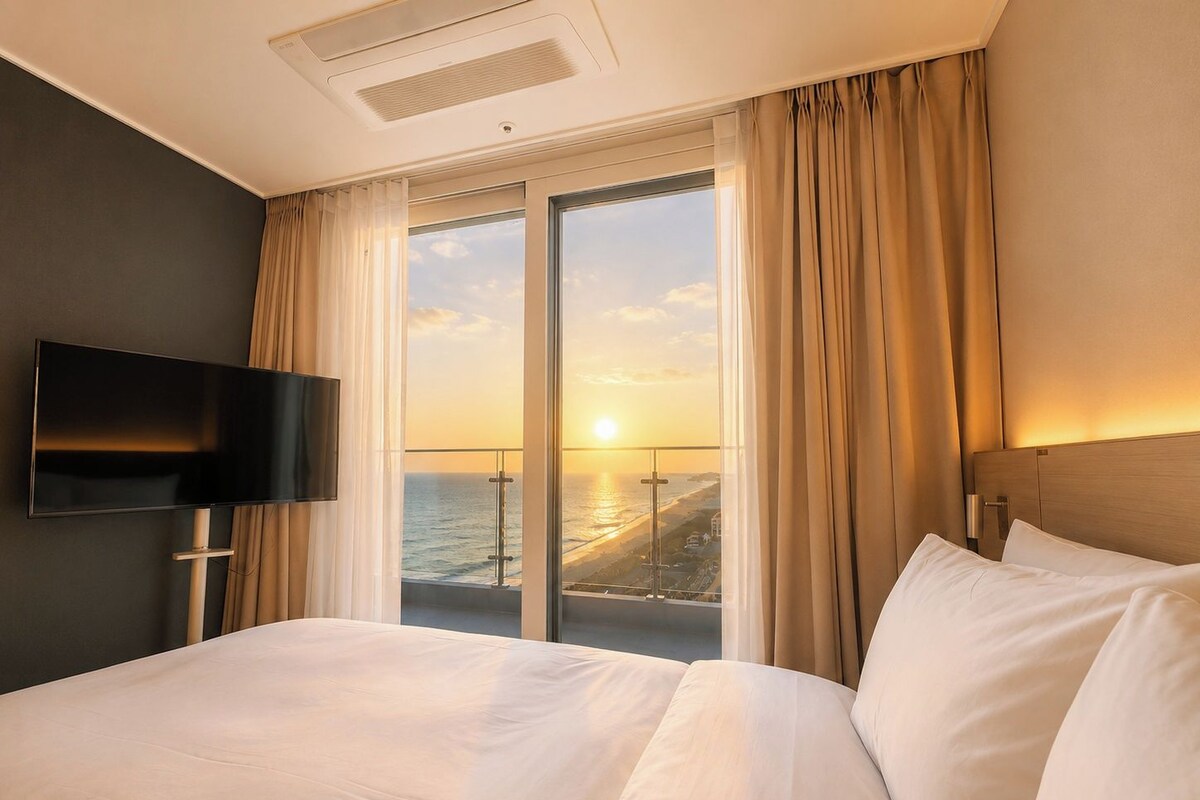 A hotel room with a bed, a television, a large window with curtains, and a view of the ocean.
