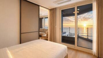 A hotel room with a bed, a seating area with a view of the sea, and a balcony with a sunset view.