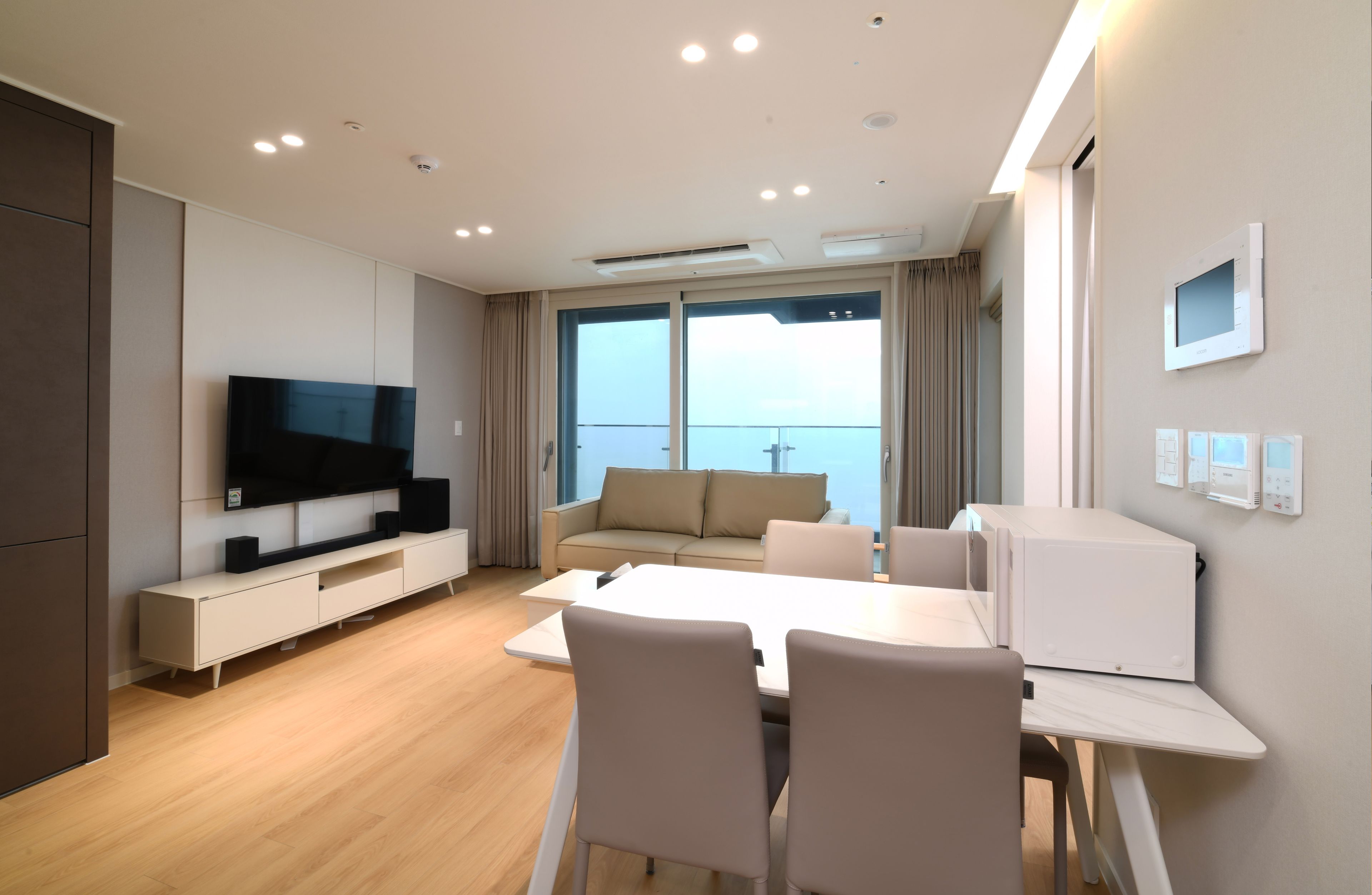 A modern hotel room with a flat-screen TV, a sofa, a dining table with chairs, and a view of the sea through large windows.