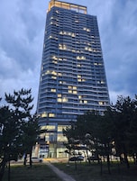 A tall, modern building with multiple floors and balconies, surrounded by trees and a parking area.