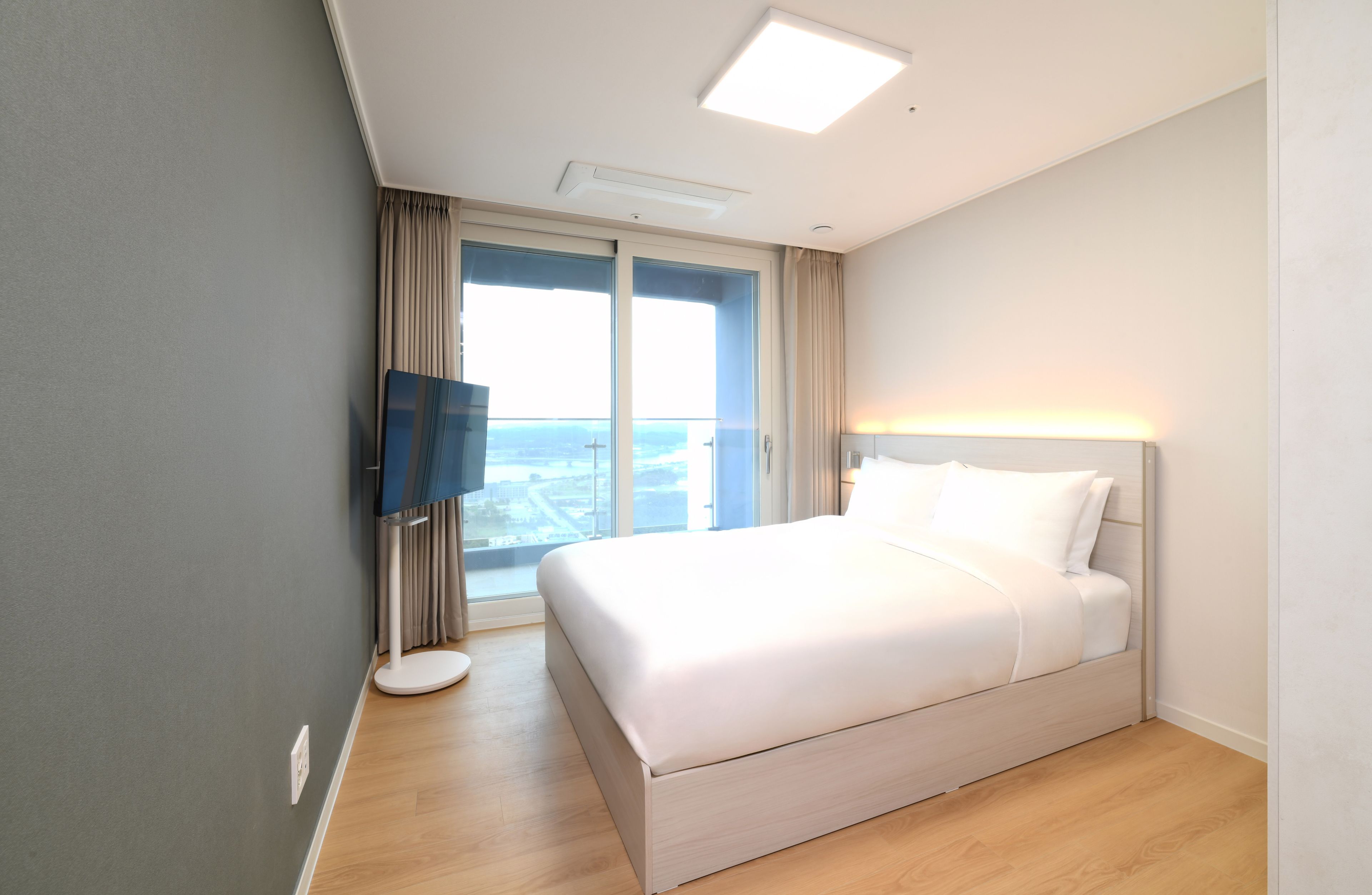 A hotel room with a bed, a television, a window with curtains, and a view of the city.