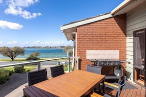 A balcony with a wooden table and chairs, a barbecue grill, and a view of a body of water.