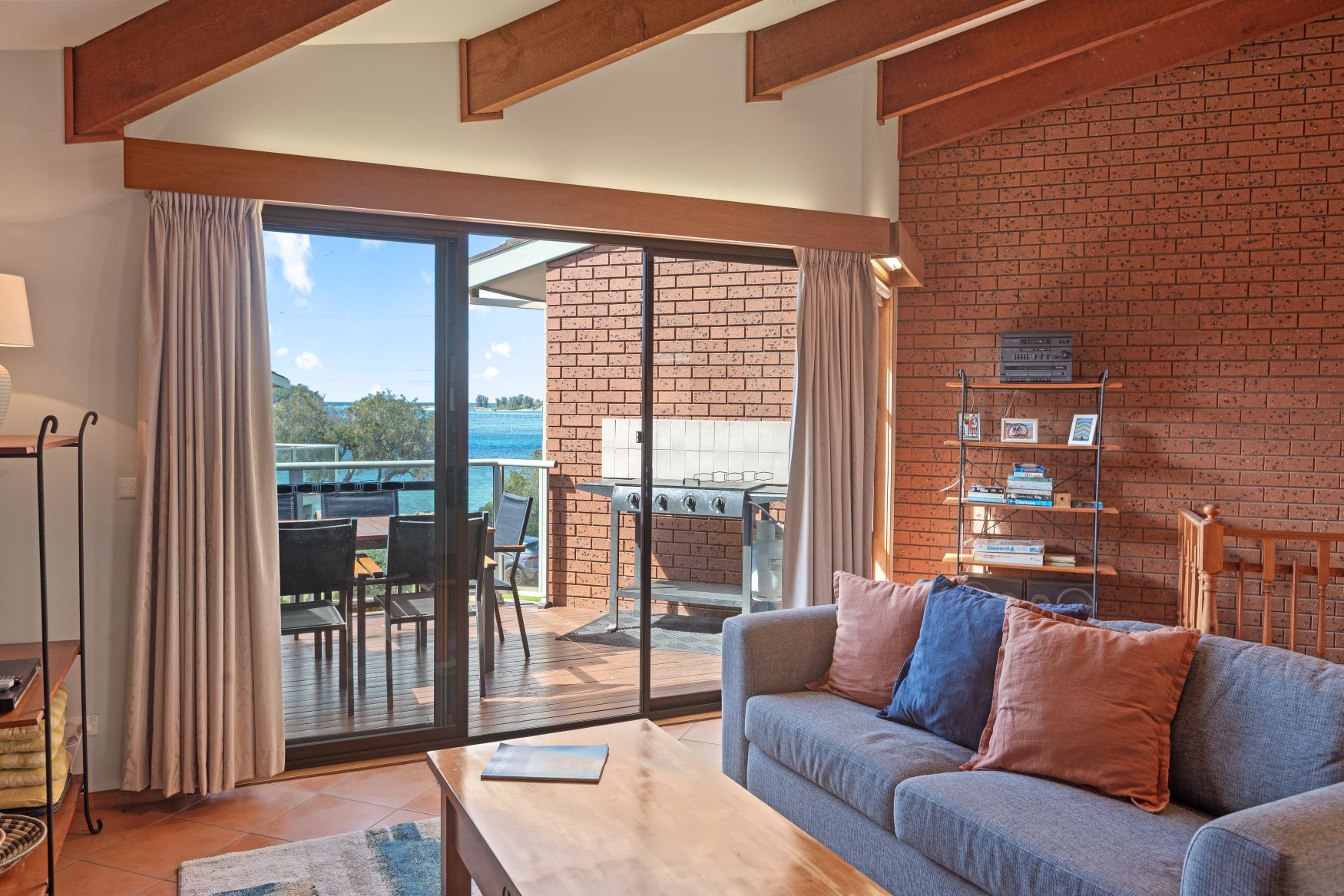 A living room with a sofa, a coffee table, and a sliding glass door leading to an outdoor patio with a grill and seating.