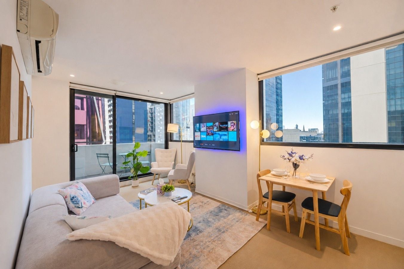 A modern living room with a sofa, a coffee table, a TV mounted on the wall, and a dining area with a table and chairs.