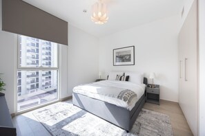 Ein modernes Schlafzimmer mit einem großen Bett, Nachttischen, einem Beistelltisch, einem gerahmten Bild an der Wand und Blick auf ein Gebäude durch das Fenster.