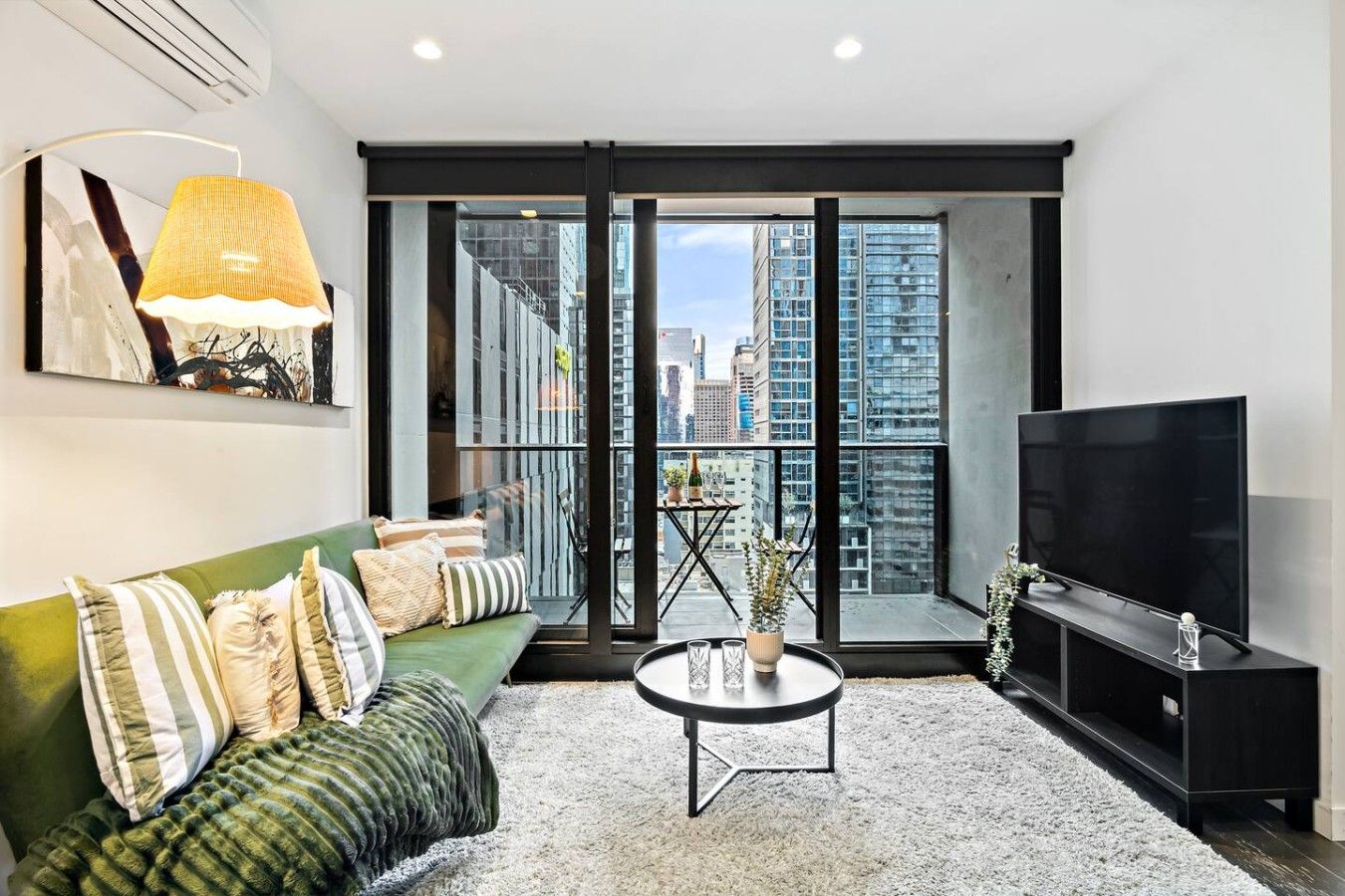 A modern living room with a green sofa, a coffee table, a flat-screen TV, and a large window offering a city view.