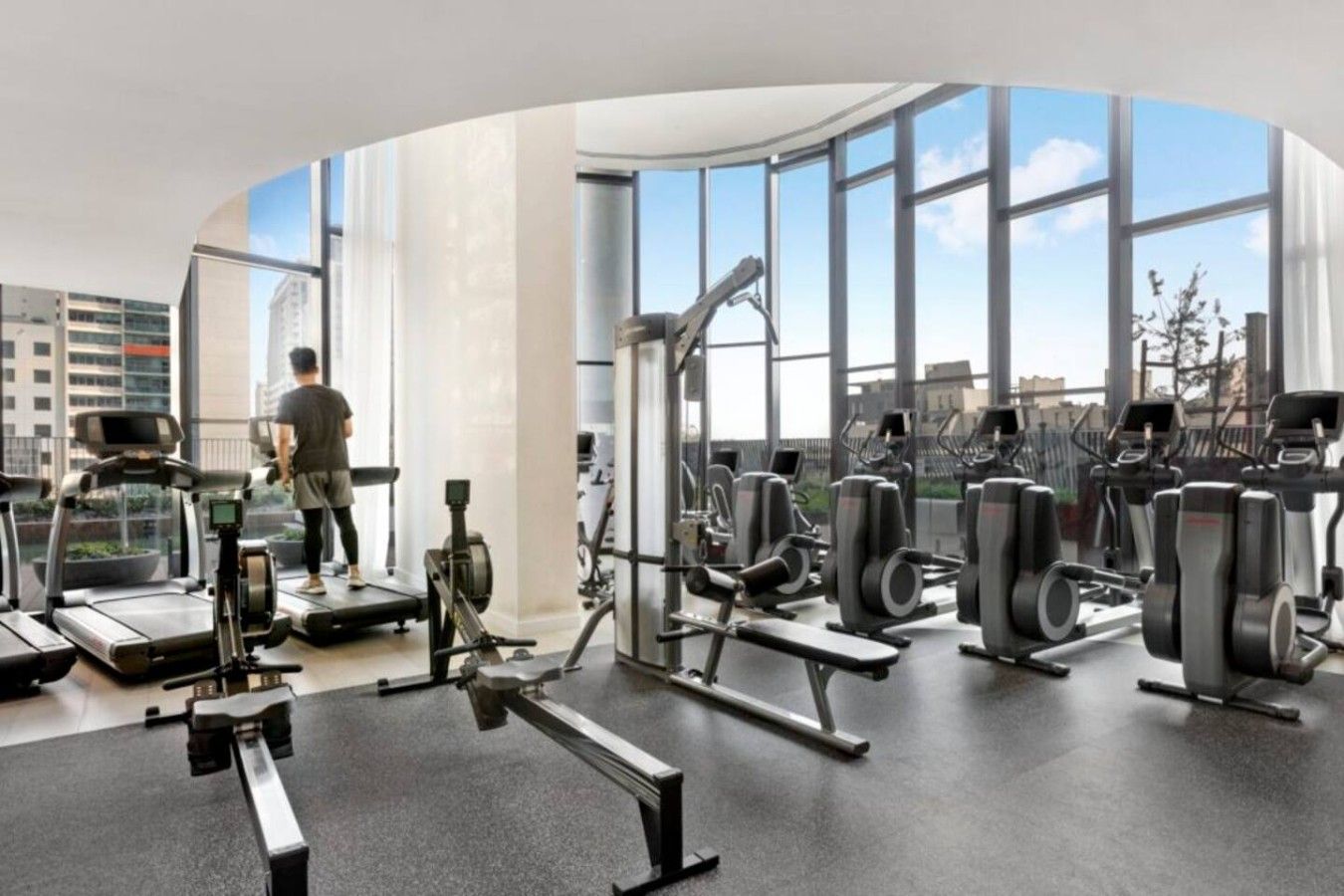 Een moderne sportschool met diverse fitnessapparaten, grote ramen met uitzicht op de stad, en een persoon die op een loopband aan het trainen is.
