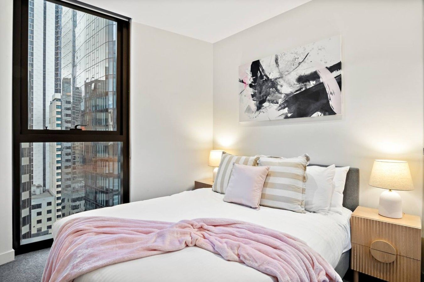 A hotel room with a large window, a bed with a pink blanket, two bedside lamps, and a black and white abstract painting on the wall.
