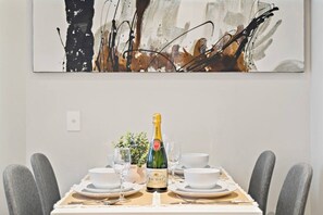 A dining table set with a bottle of champagne, glasses, and plates, with a modern abstract painting above. - Elegant 2BR Apt | Panoramic Views • Pool & Gym (Melbourne)