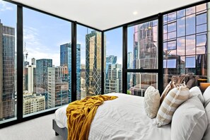 A bedroom with a large bed, a yellow throw blanket, and a view of a cityscape through floor-to-ceiling windows. - Cozy 2BR Apt | Vibrant Living w/ Pool & Gym (Melbourne)