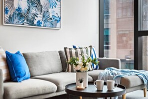 Una sala moderna con un sofá gris, cojines azules y a rayas, una mesita redonda con un jarrón blanco y dos tazas blancas, y un amplio ventanal con persianas.
