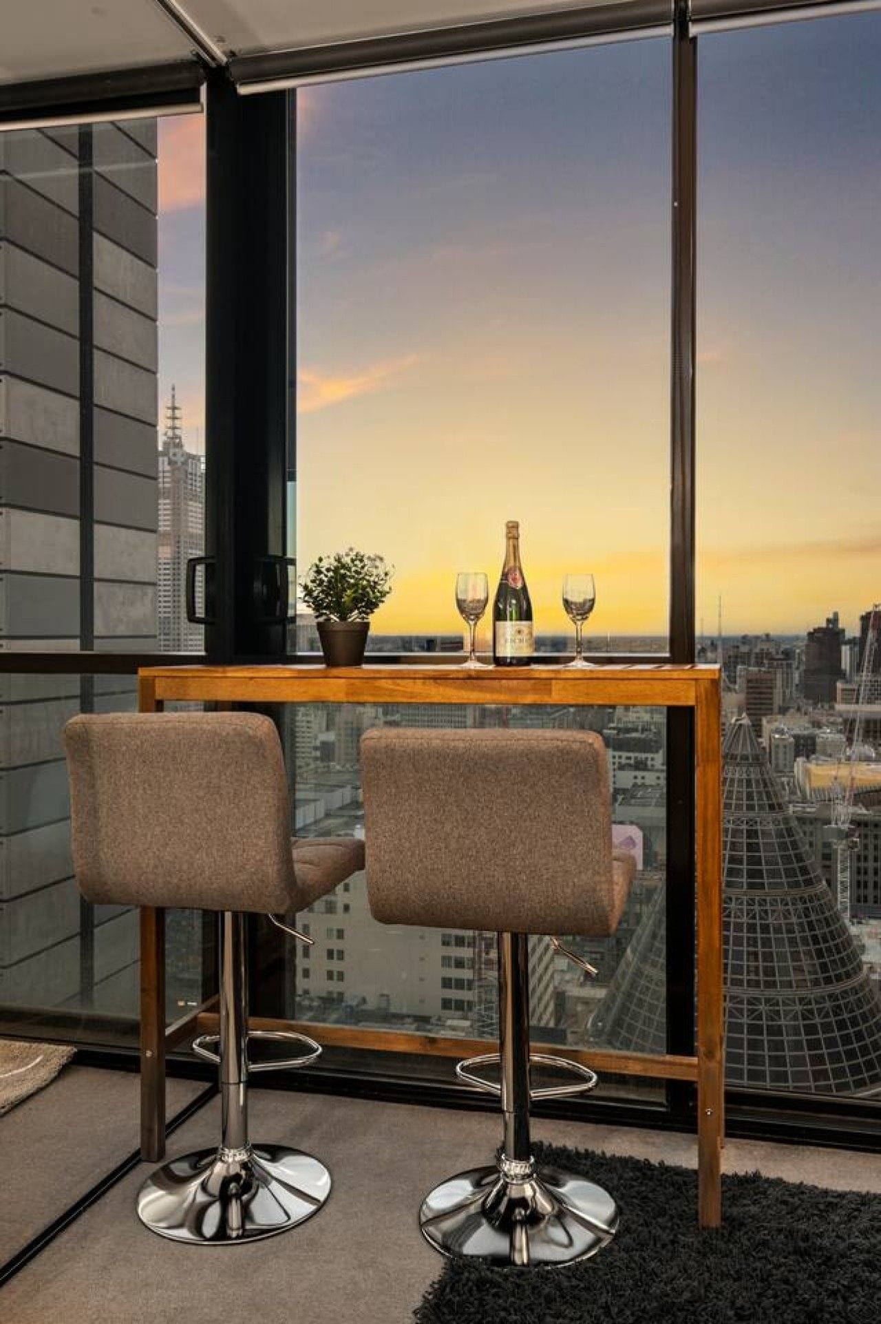 A high-rise room with a city view, a wooden table with two bar stools, a bottle of champagne, and two glasses.