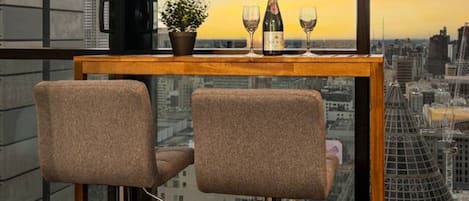 A high-rise room with a city view, a wooden table with two bar stools, a bottle of champagne, and two glasses.