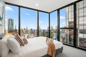 Ein modernes Hotelzimmer mit einem großen Bett, vielen Kissen und Blick auf eine Stadtlandschaft durch raumhohe Fenster.