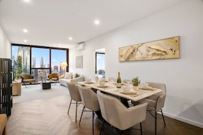 A modern dining area with a city view, a large painting on the wall, and a table set for a meal. - LVL 56 City Skyline | Stylish 3B Apt w/ Pool & Gym (Melbourne)