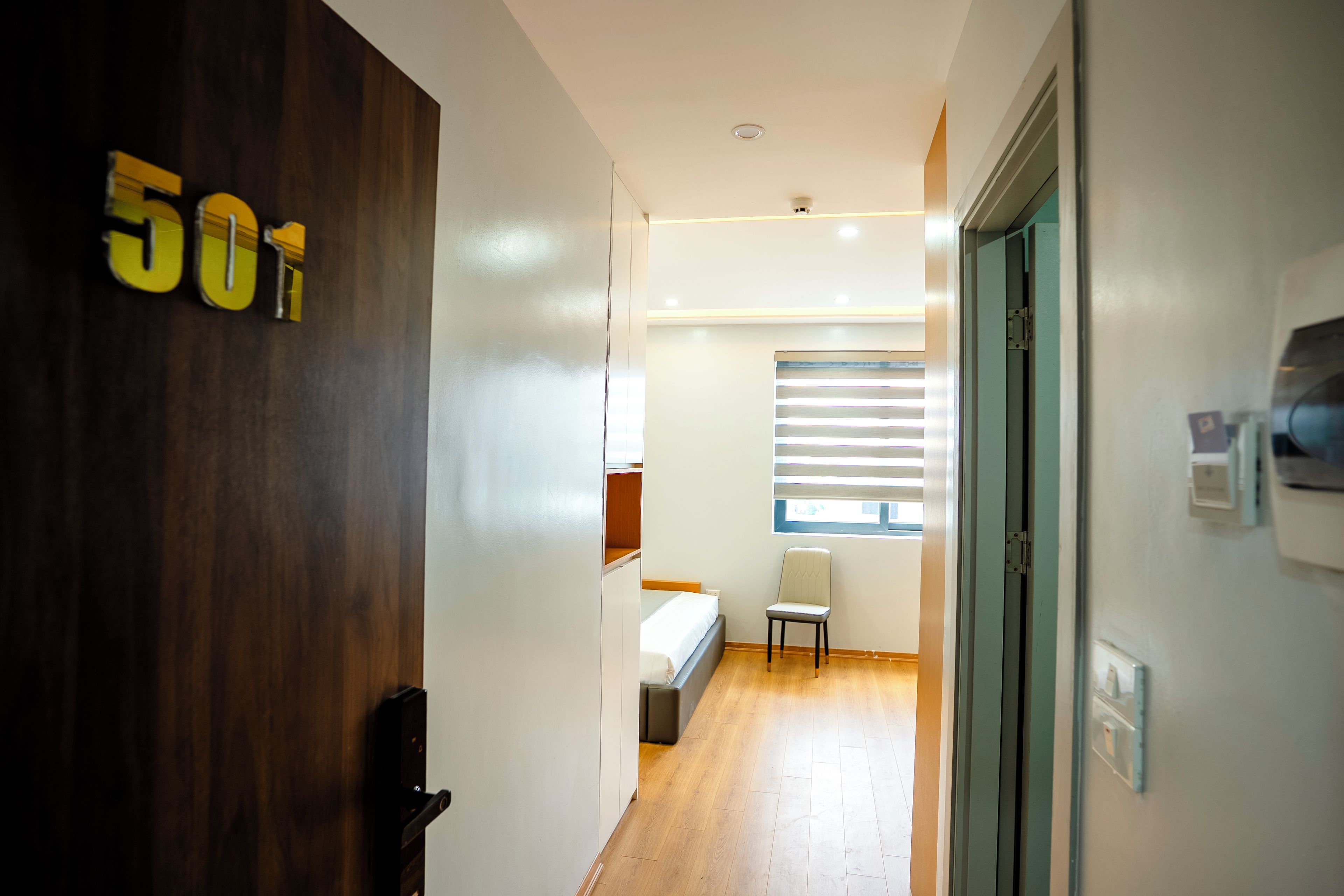 Pasillo de hotel con puerta de madera identificada con el número '501', una cama, una silla y un ventanal con persianas.