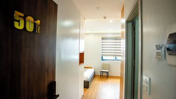 Pasillo de hotel con puerta de madera identificada con el número '501', una cama, una silla y un ventanal con persianas.