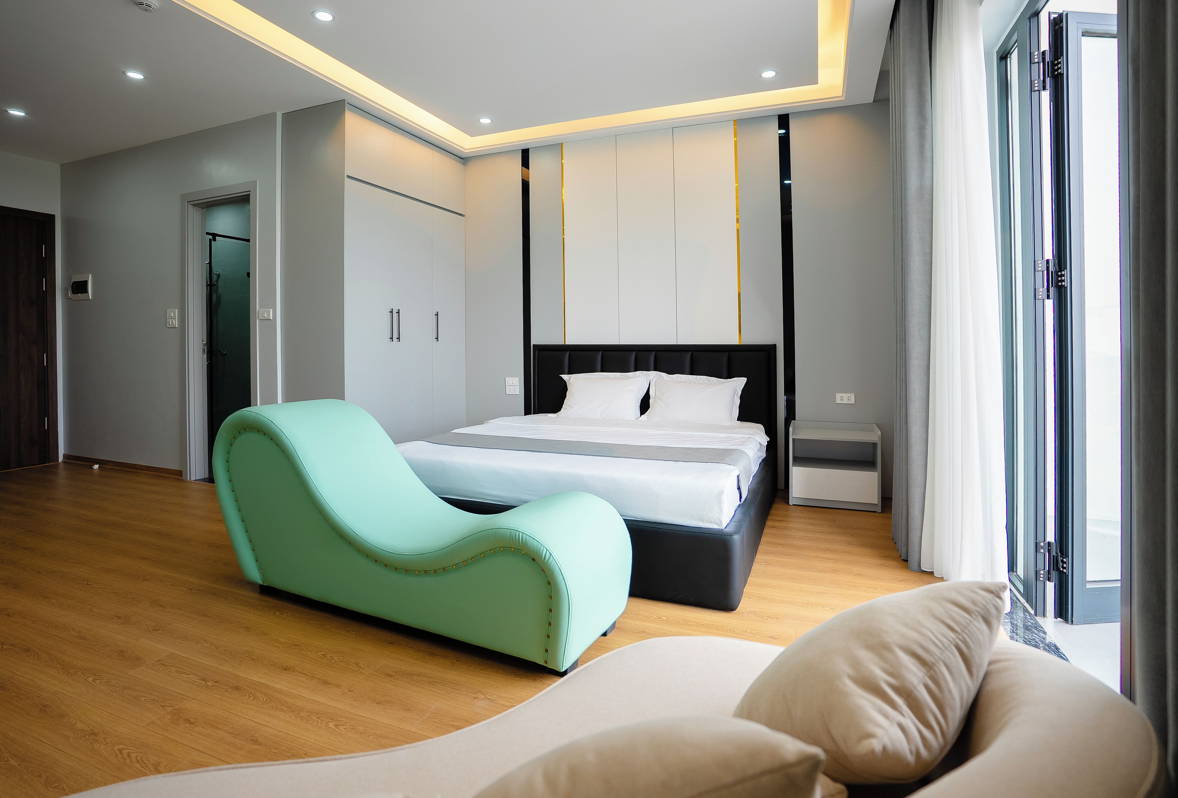 Un dormitorio moderno con una cama, un sillón curvo color turquesa y un ventanal grande.
