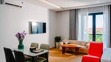 A modern living room with a dining area, a flat-screen TV mounted on the wall, a sofa, a red chair, and a balcony with a view.