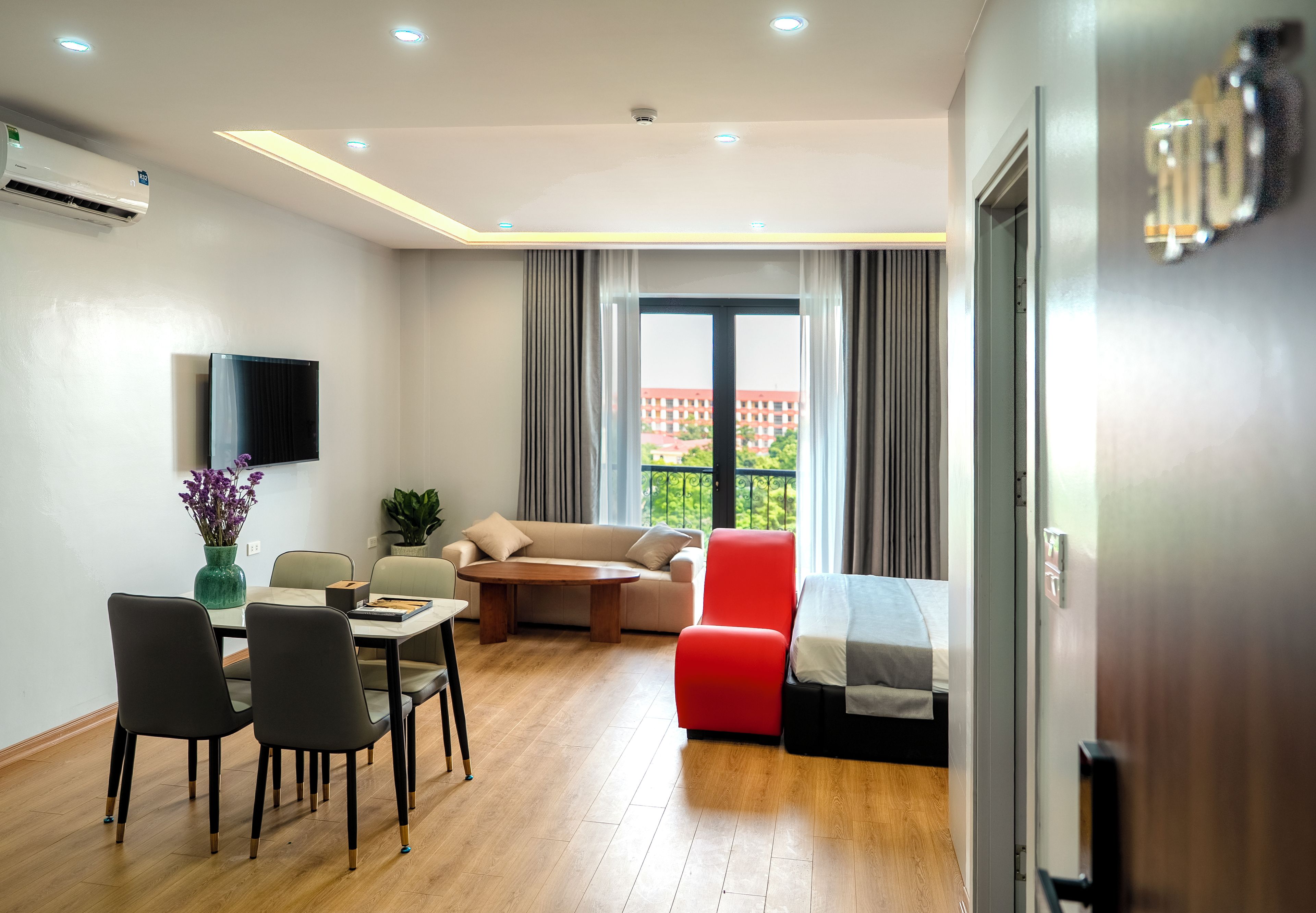 A modern hotel room with a dining area, a sofa, a red chair, a bed, and a large window with a view of buildings.