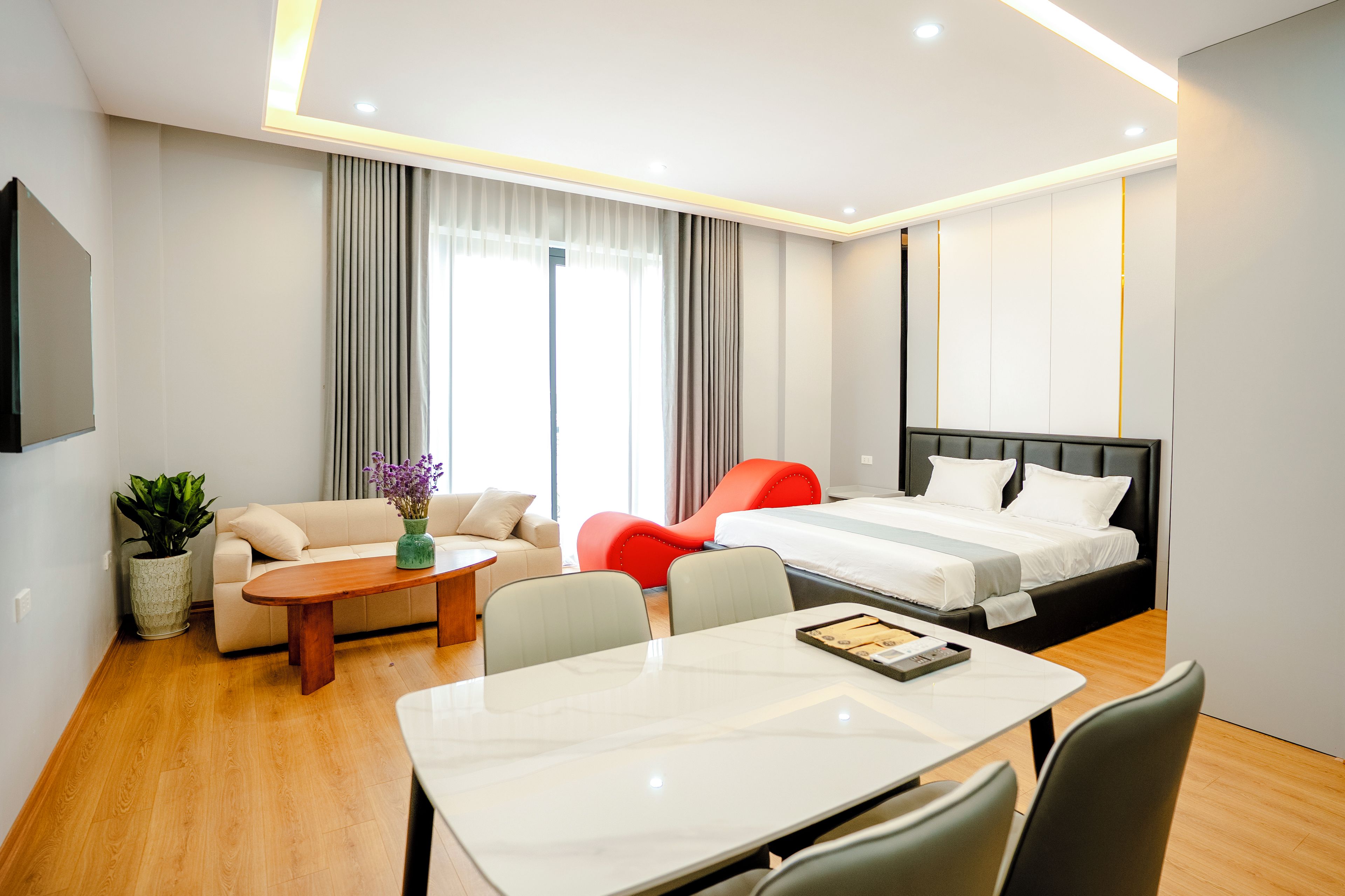 A modern hotel room with a bed, a sofa, a dining table, and a small table with a vase of flowers.