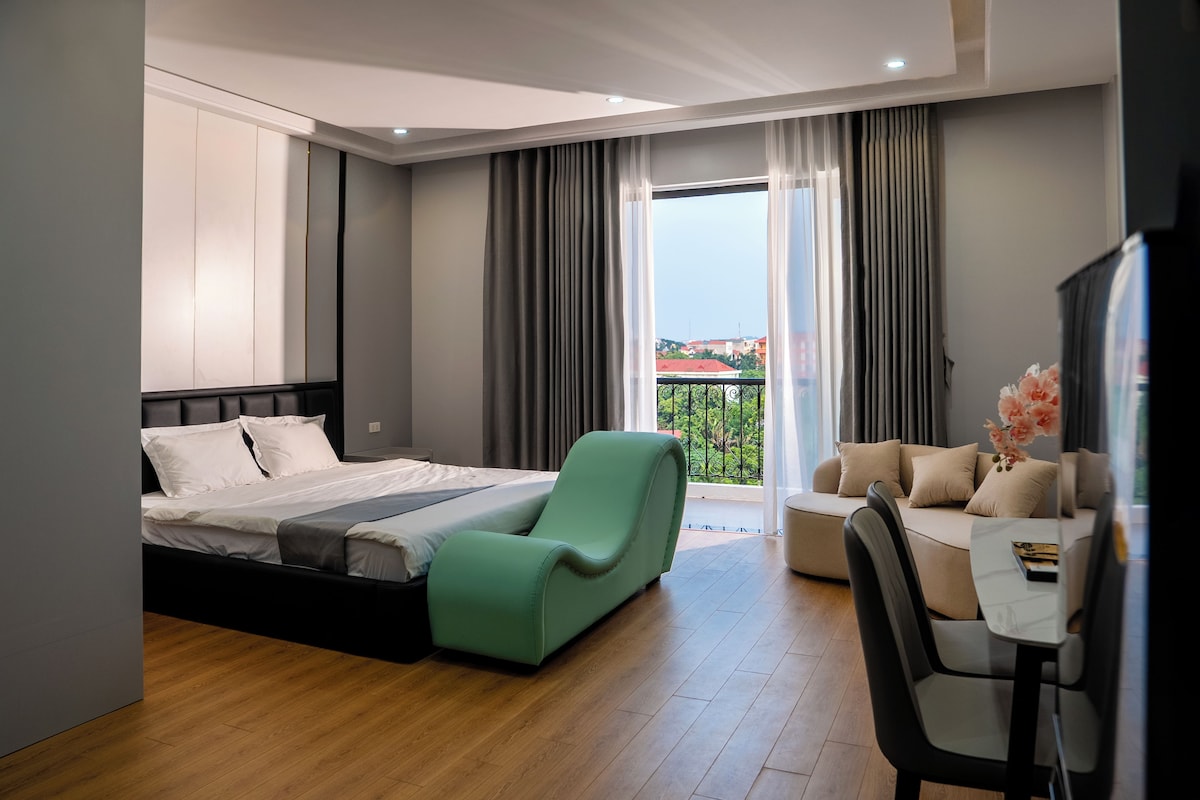 Habitación de hotel moderna con cama, un sillón verde, sofá, mesa de comedor y balcón con vistas.